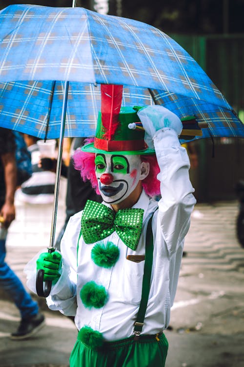 Free Anonymous male clown in funny costume and pink wig walking on street with umbrella and looking at camera Stock Photo