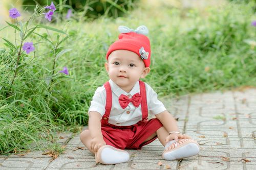 Free Adorable little boy in red shorts bow tie and hat sitting on ground with grass and flowers behind Stock Photo