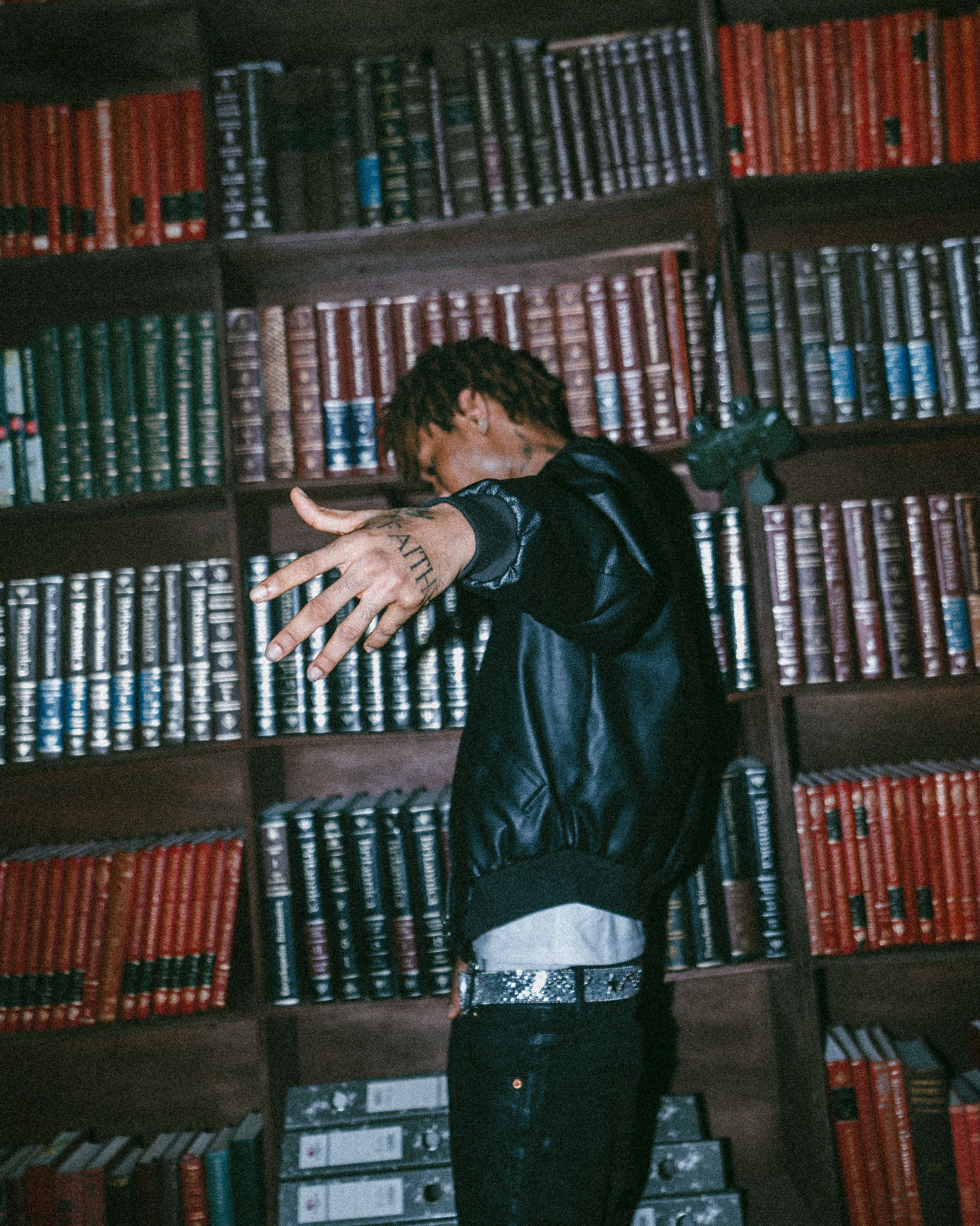 A young man in a leather jacket extends his arm in front of bookshelves.