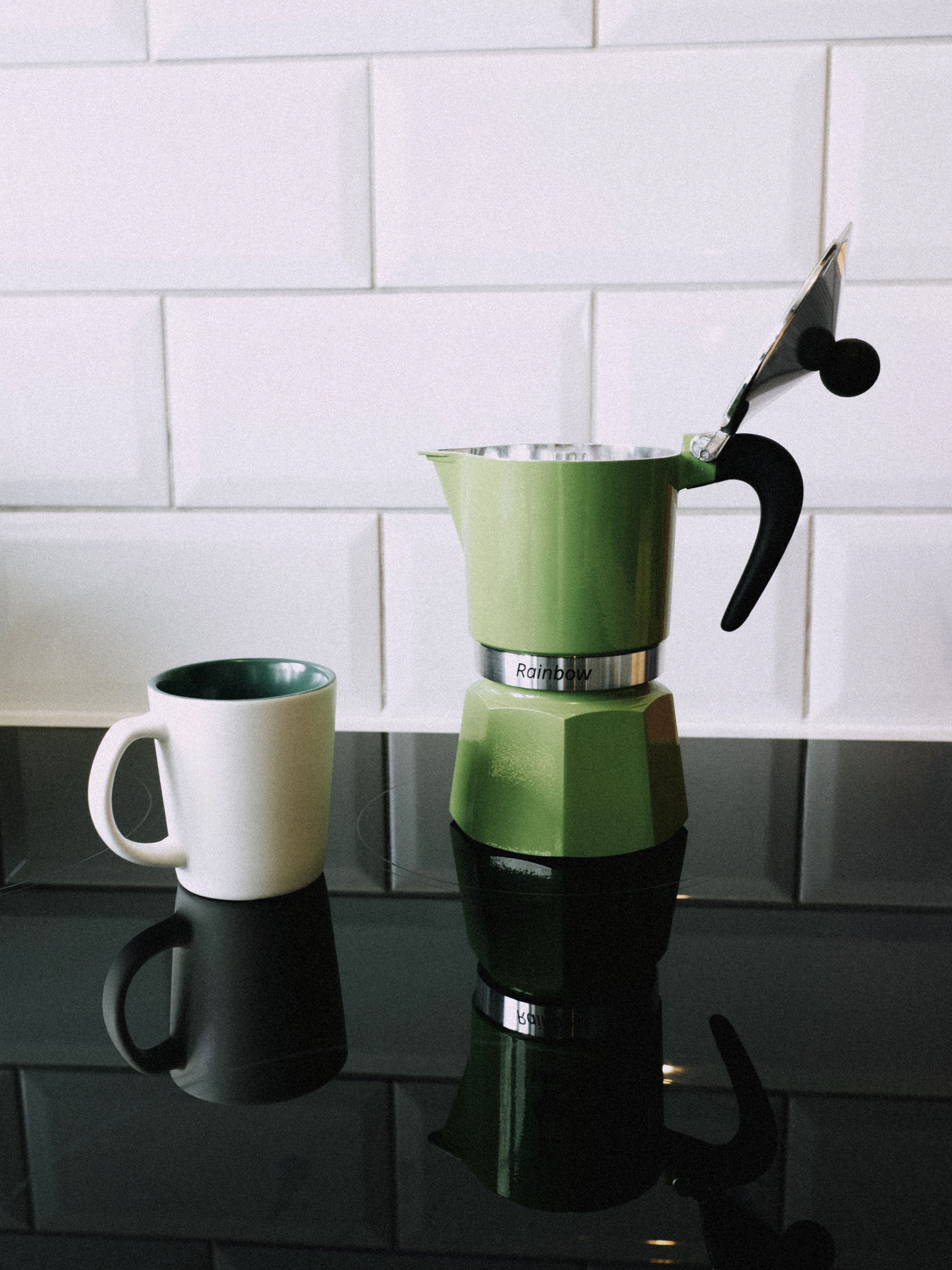 A stylish green moka pot and mug on a sleek black countertop with white tile backdrop.
