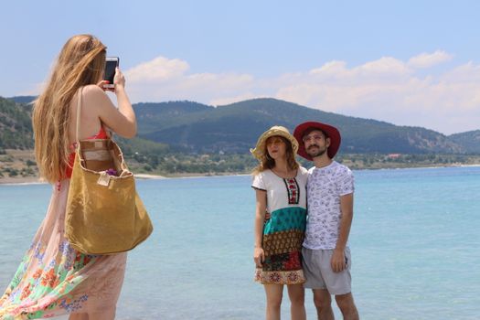 A woman photographs a couple at a picturesque beach with mountains in the background.