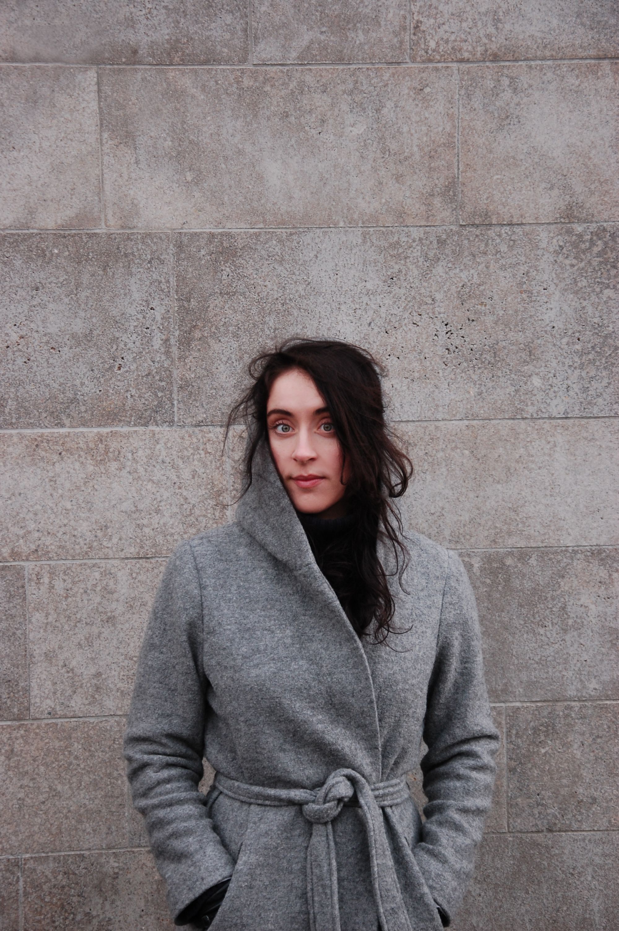 Woman in a grey coat posing confidently against a stone wall, chilly weather.