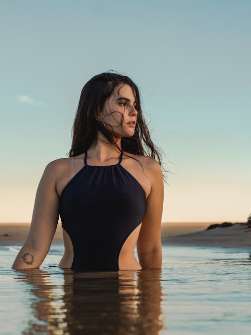 Free Gentle brunette in black swimsuit standing waist deep in water and looking away in sunset Stock Photo