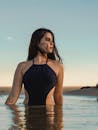 穿着黑色泳衣的温和黑发女子站在齐腰深的水中，在日落中目光移开