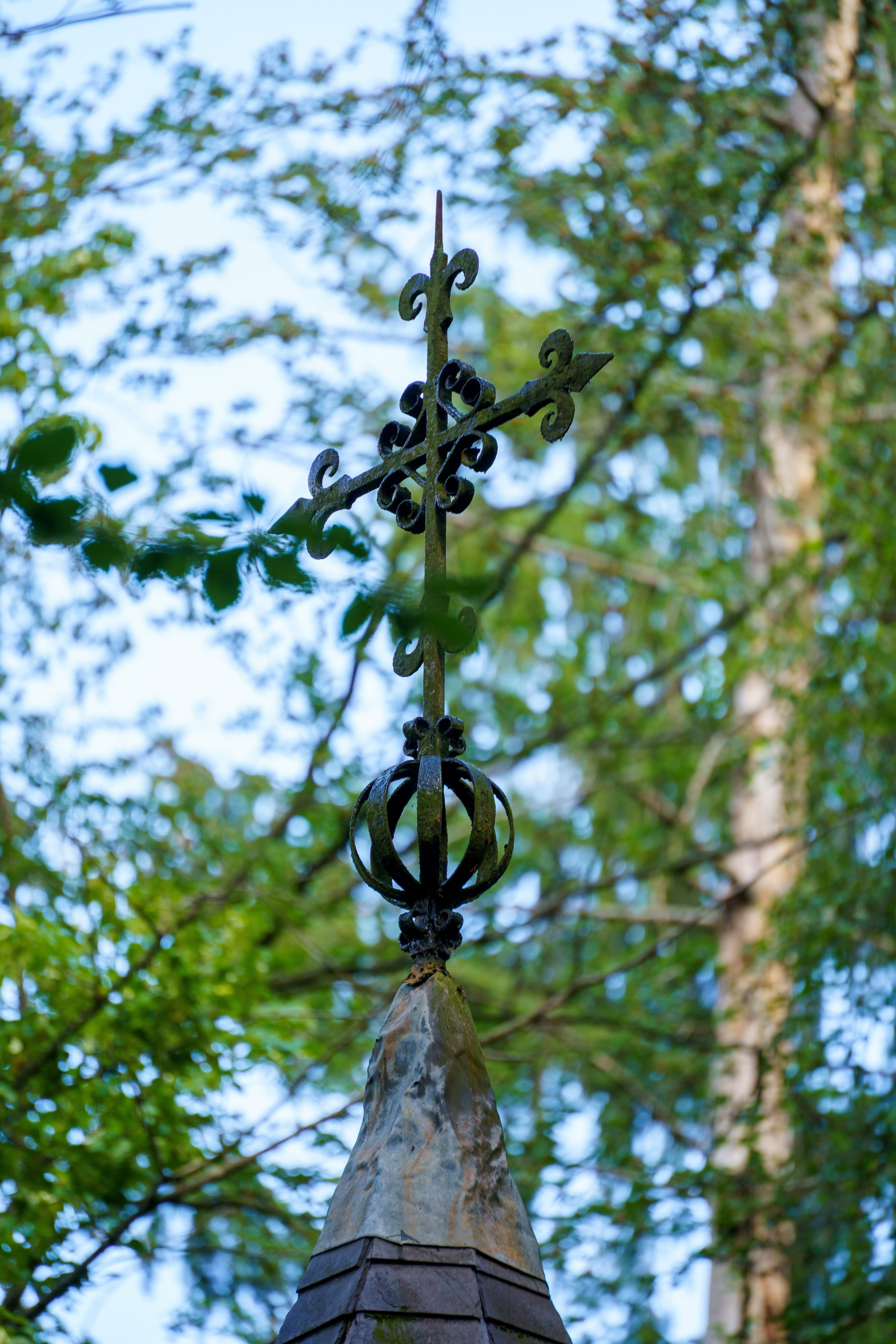 Ornate gothic iron cross atop a spire amidst lush greenery in Vorselaar, Belgium.