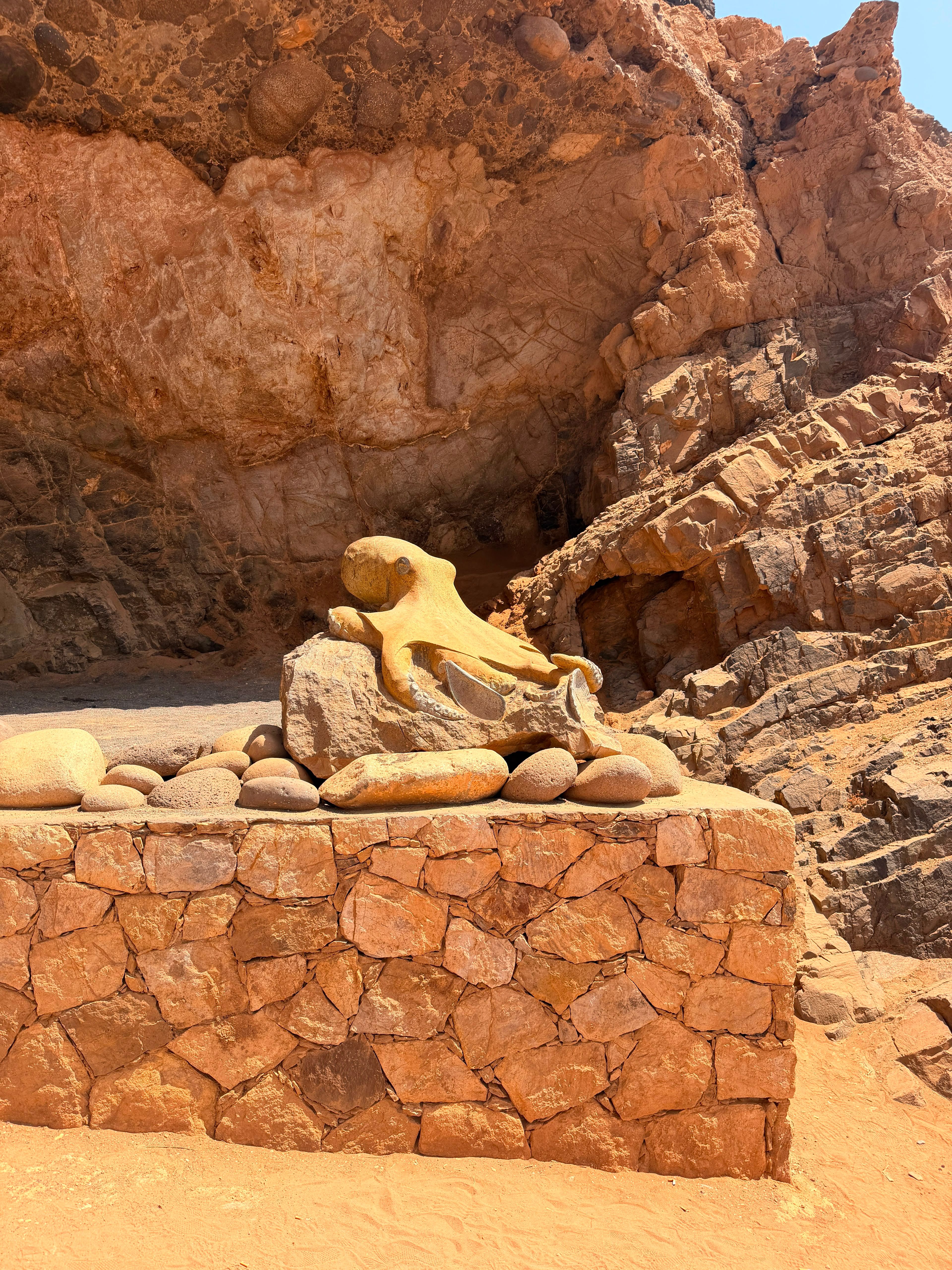 Intricate octopus sculpture rests on stone wall in arid desert setting.