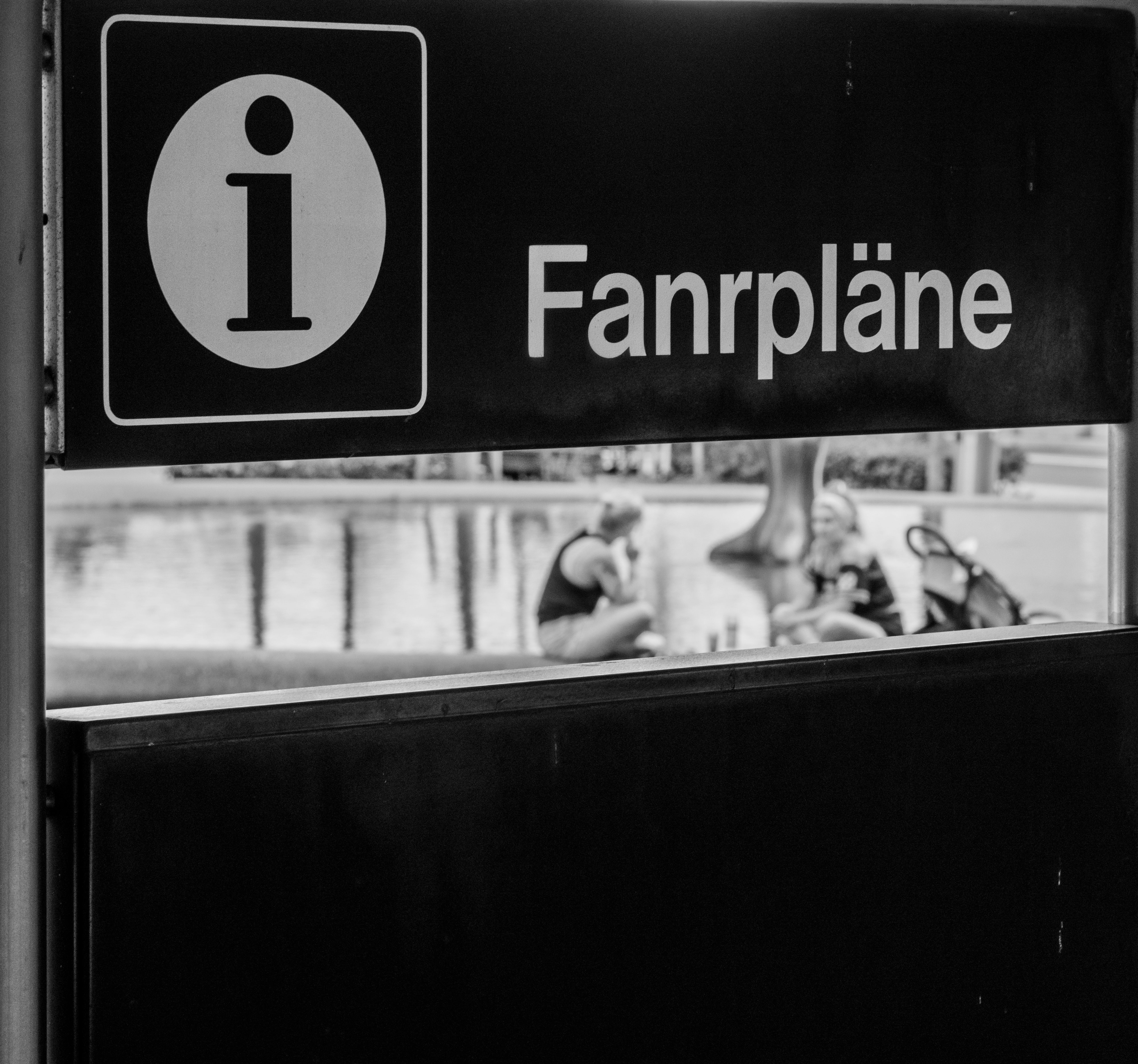 Monochrome photo of an information sign and people by a fountain in Basel, Switzerland.