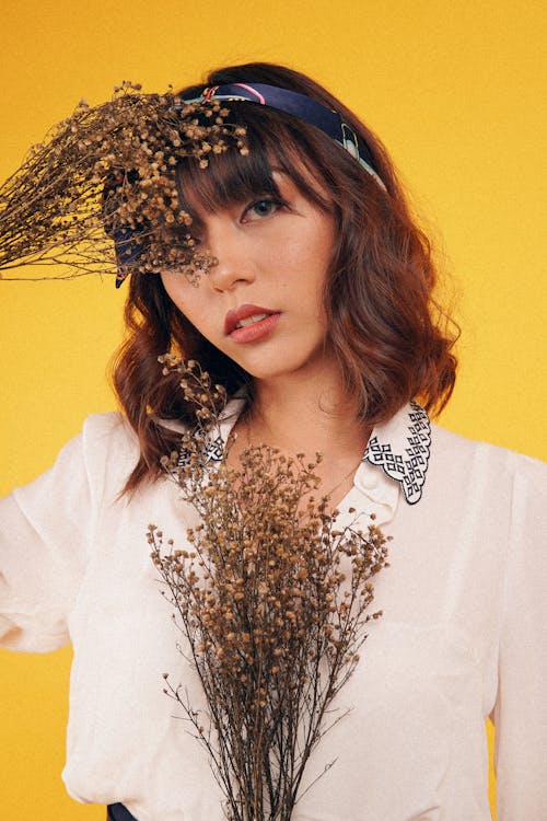 Free Young woman in light white blouse covering eye with dry bouquets looking at camera on yellow background Stock Photo