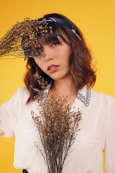 一位穿着浅色衬衫的年轻女子用干花束遮住眼睛，在黄色背景前看向相机