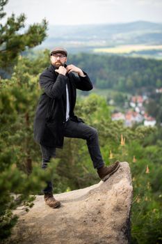 Man in stylish attire striking a pose atop a rock, with stunning views of Jonsdorf, Germany.