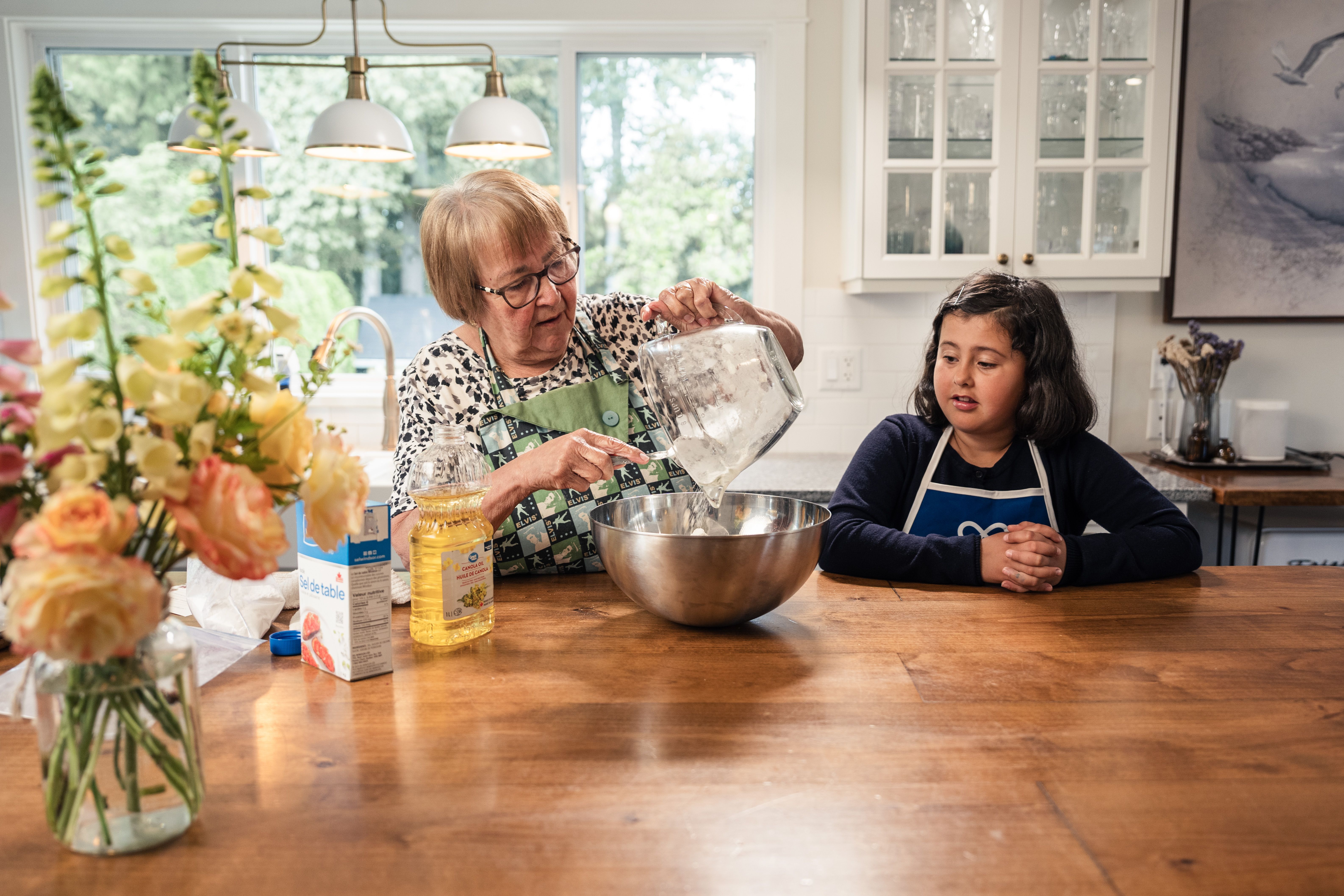 A grandmother and granddaughter baking in a kitchen filled with natural light and flowers.