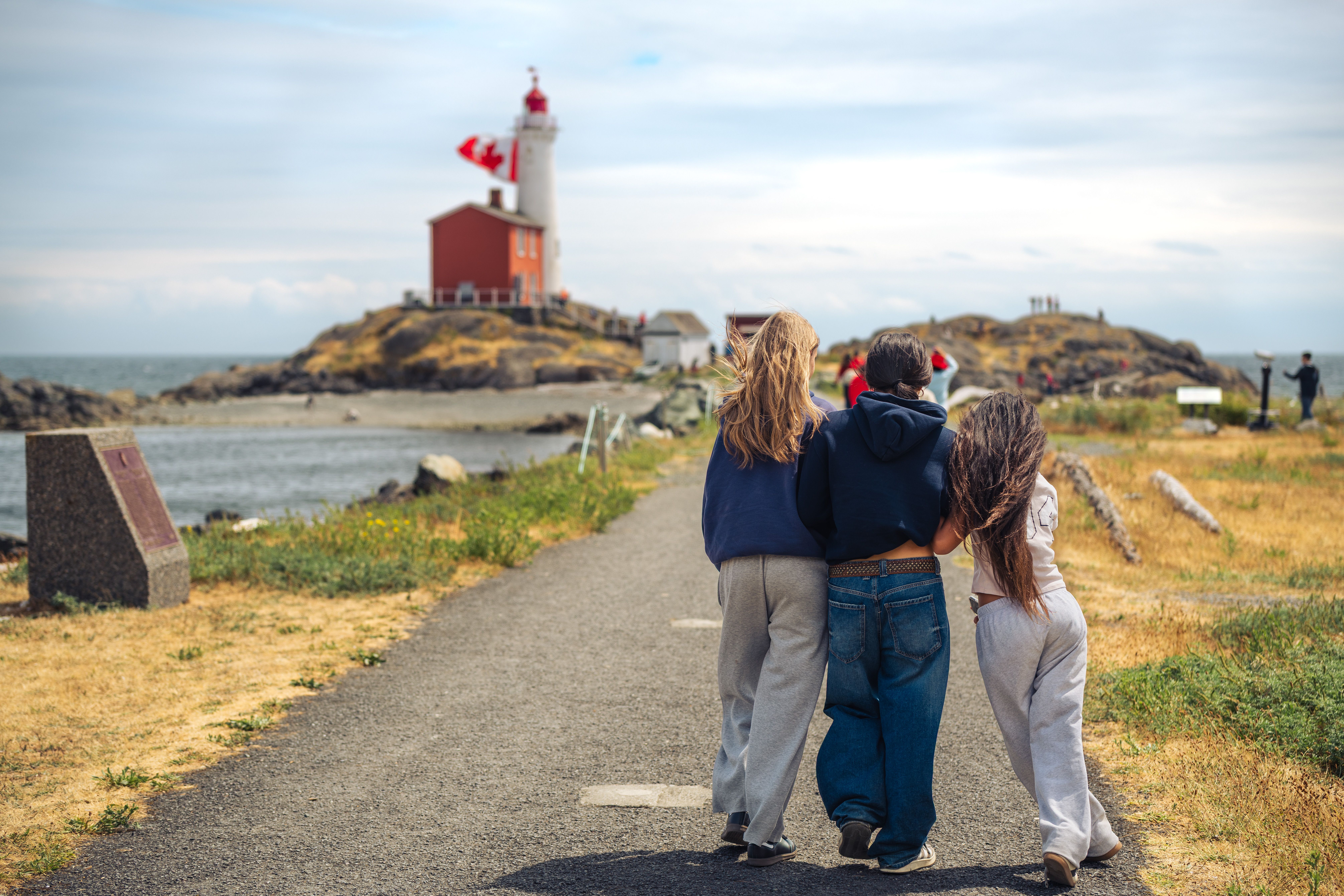 Three friends walk towards a lighthouse on a sunny day in Victoria, British Columbia, Canada.