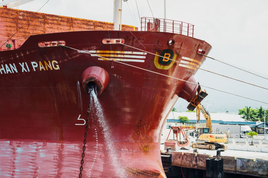 Front view of a large red cargo ship docked, releasing water, with construction equipment nearby.