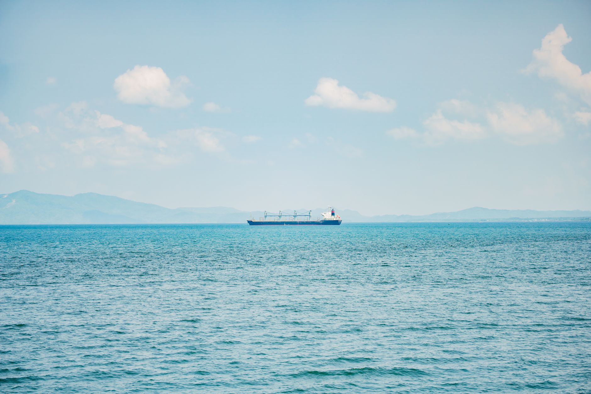 Illustrative photograph: A serene view of a cargo ship sailing on a calm ocean with distant mountains and clear sky.