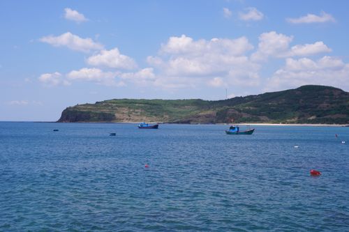 Free Peaceful coastal landscape featuring fishing boats, calm blue waters, and lush green hills under a bright sky. Stock Photo