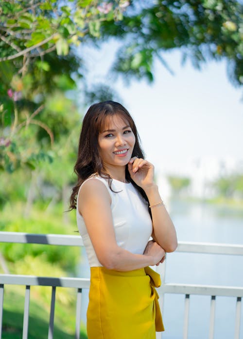 Content modern ethnic female in multicolored dress smiling while standing at fence of bridge in summer park