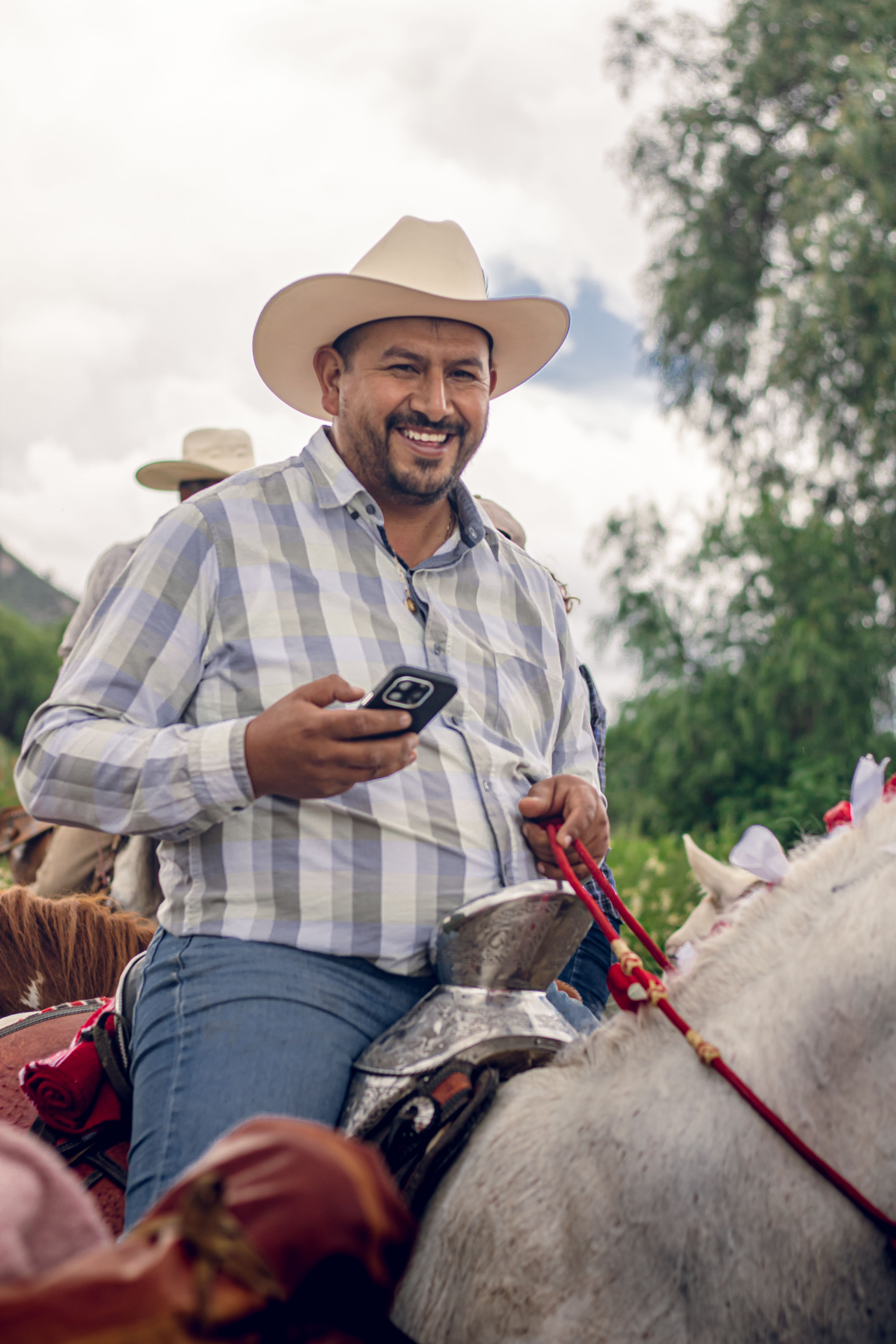 A smiling Mexican cowboy rides a horse while using a smartphone, showcasing modern and traditional lifestyles in Hidalgo.