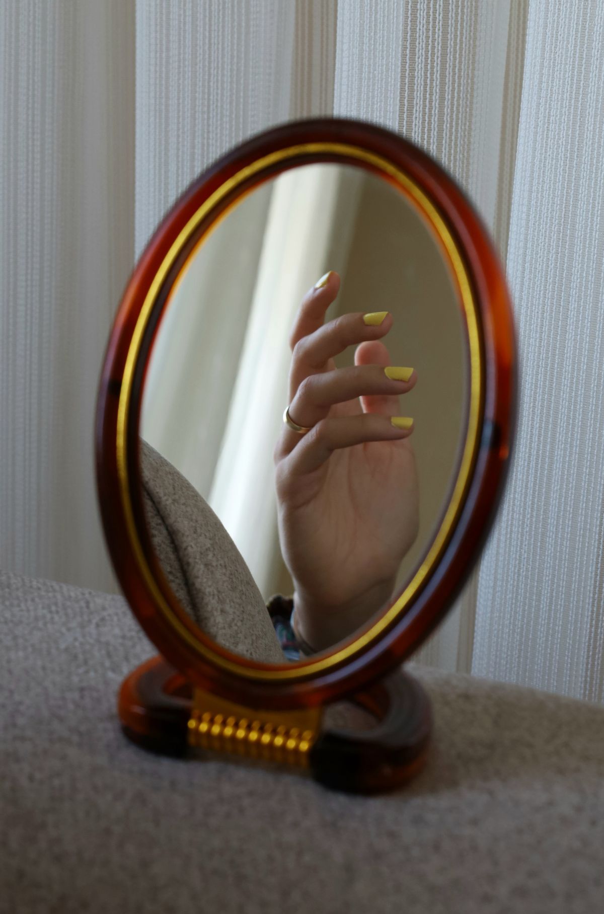 A hand with yellow polished nails reflected in an oval mirror indoors.