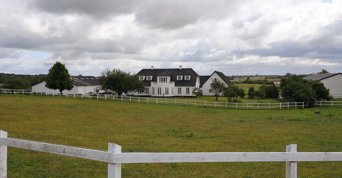 A scenic view of a traditional Danish ranch with white fencing and lush greenery under a cloudy sky.