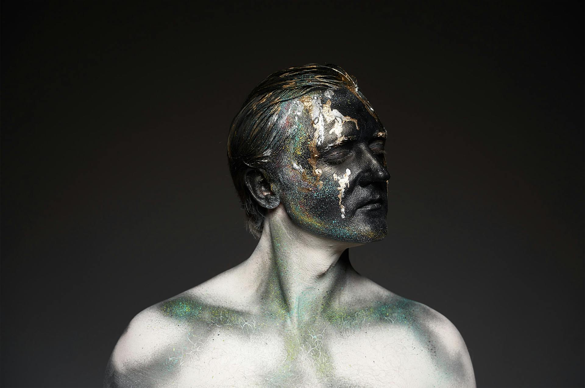 Male shirtless model with glowing glitters on black painted face covering eyes standing on black backdrop