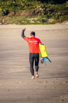 A surfer in a red wetsuit walks on a sandy beach carrying a yellow bodyboard, ready for action.