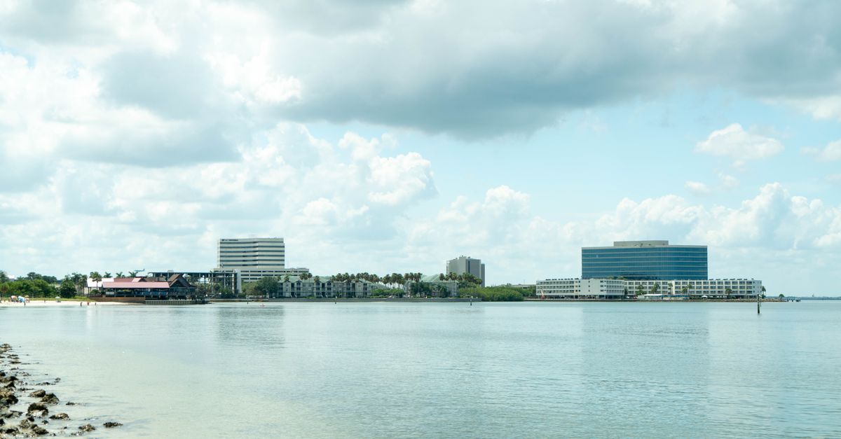 Coastal urban landscape with buildings, clear skies, and calm waters reflecting clouds.