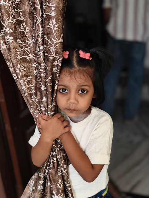 Free Portrait of a young girl with expressive eyes peering around a patterned curtain inside a home. Stock Photo