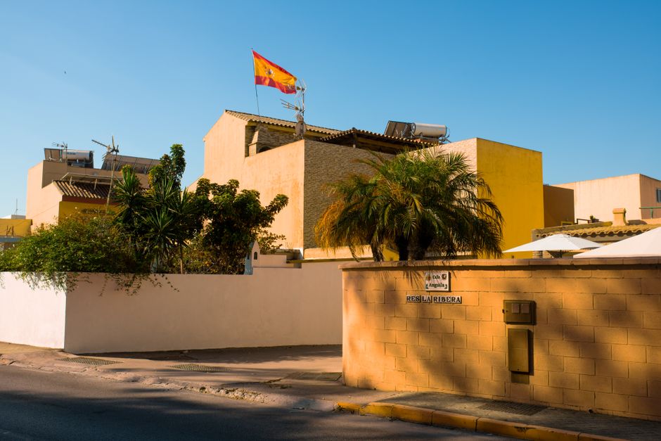 Bandera española ondeando sobre una zona residencial con edificios amarillos y vegetación frondosa.