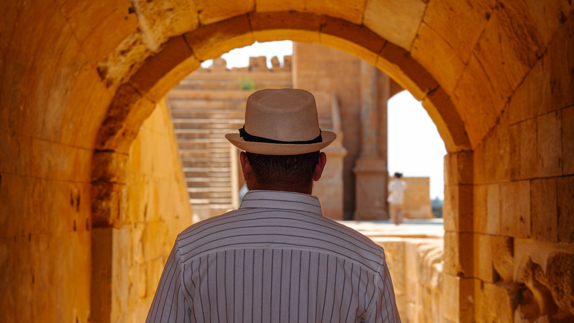 A man in a striped shirt and straw hat walking through ancient Libyan ruins under an archway.. Photo by abdo alshreef on Pexels