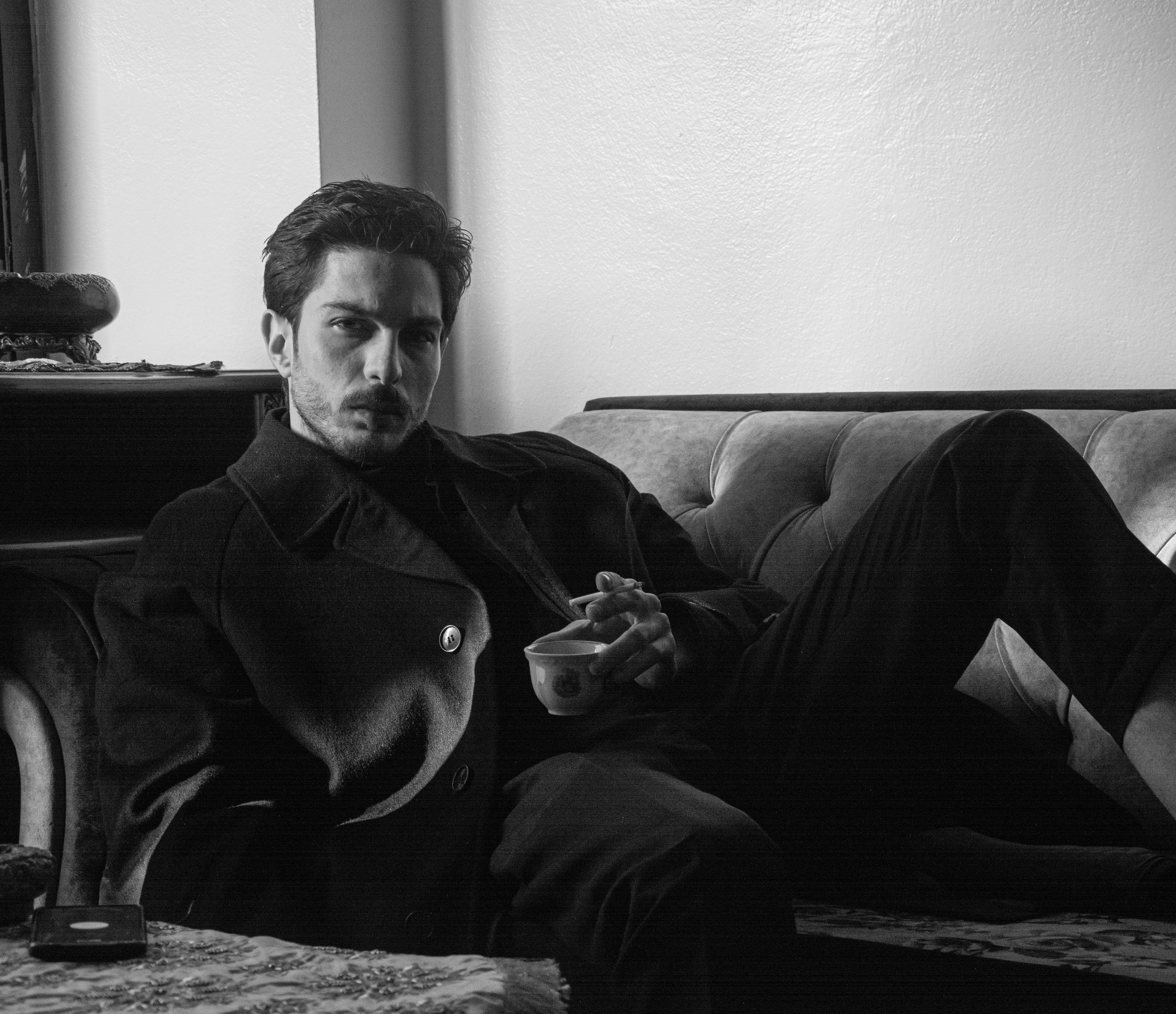 Black and white image of a man relaxing on a sofa holding a cup, creating a moody atmosphere.