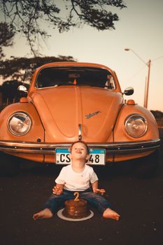 A child sits in front of an orange vintage car with a birthday cake outdoors.