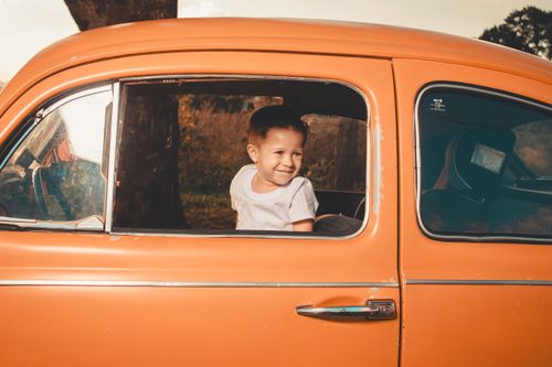 Free A cheerful child smiling while looking outside from a vintage orange car window in daylight. Stock Photo
