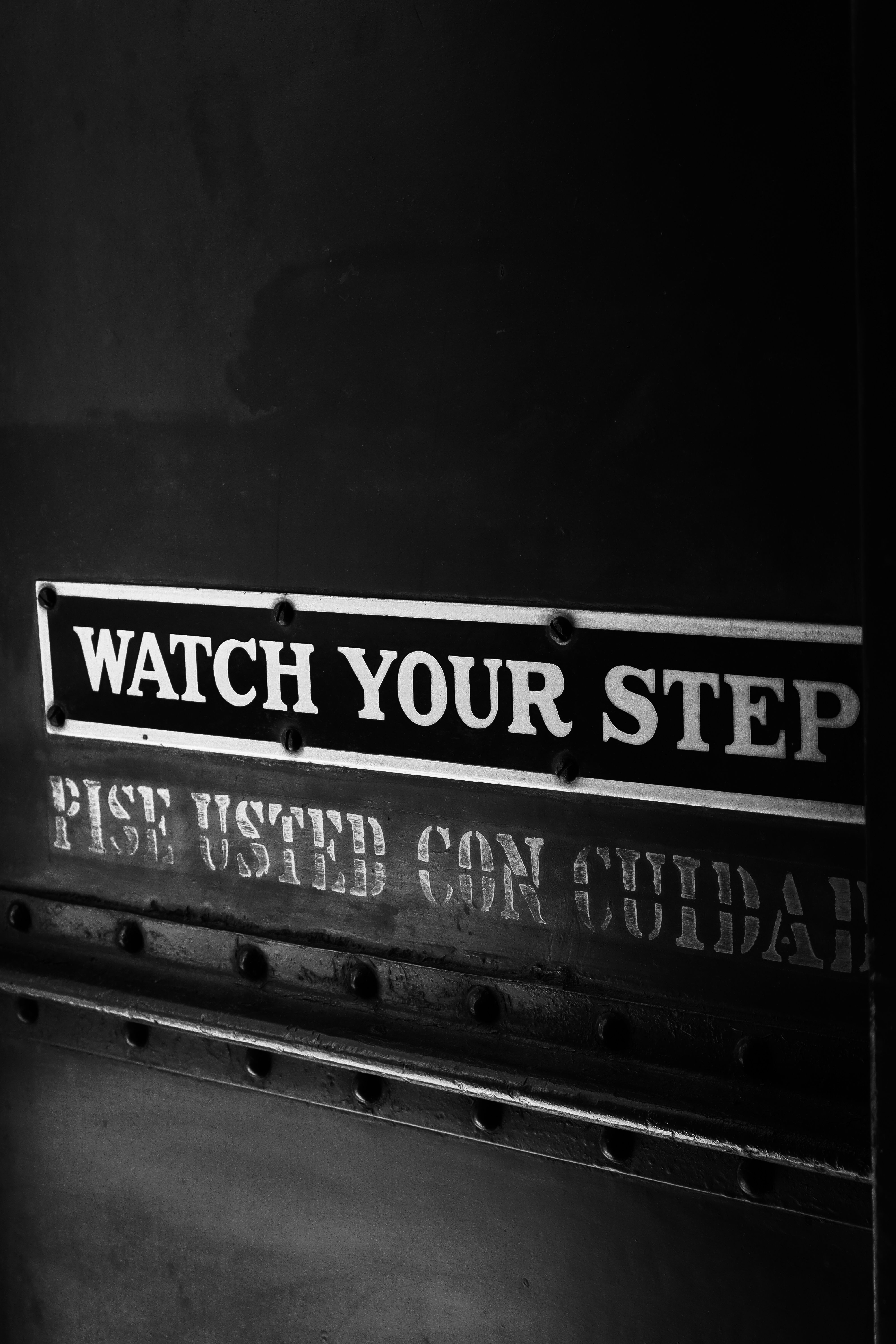 Black and white photo of a 'Watch Your Step' sign, invoking precaution.