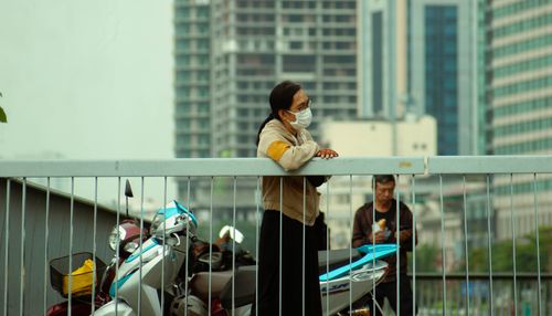 Free Two people wearing masks lean over a bridge with urban buildings in the background. Stock Photo