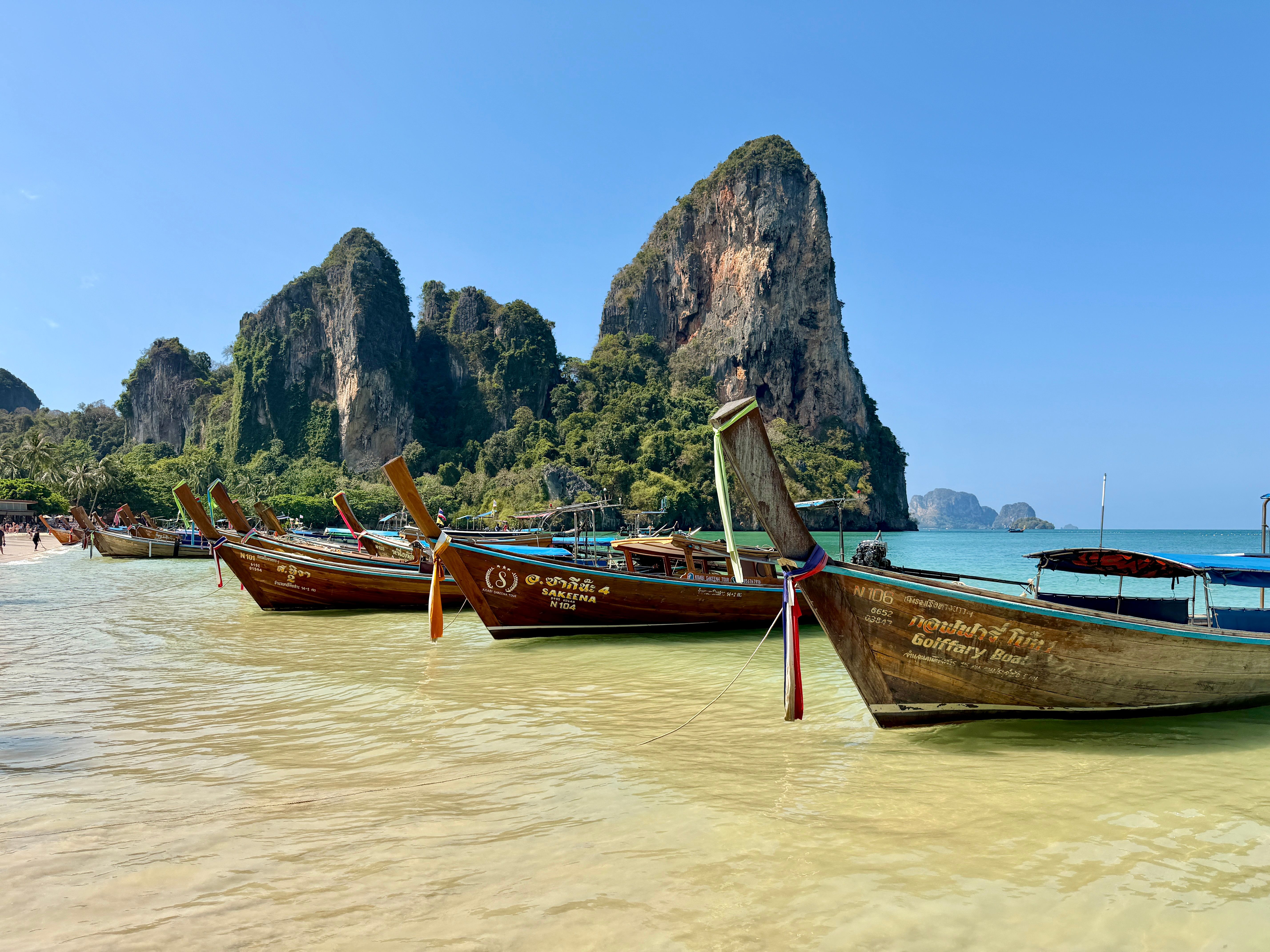 Wooden longtail boats line the beach at scenic Railay Bay, Thailand, with limestone cliffs in the background.