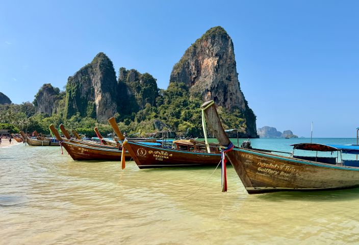 Wooden longtail boats line the beach at scenic Railay Bay, Thailand, with limestone cliffs in the background.