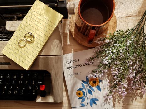 Charming vintage flatlay with a typewriter, coffee mug, flowers, and rings on a wooden surface.