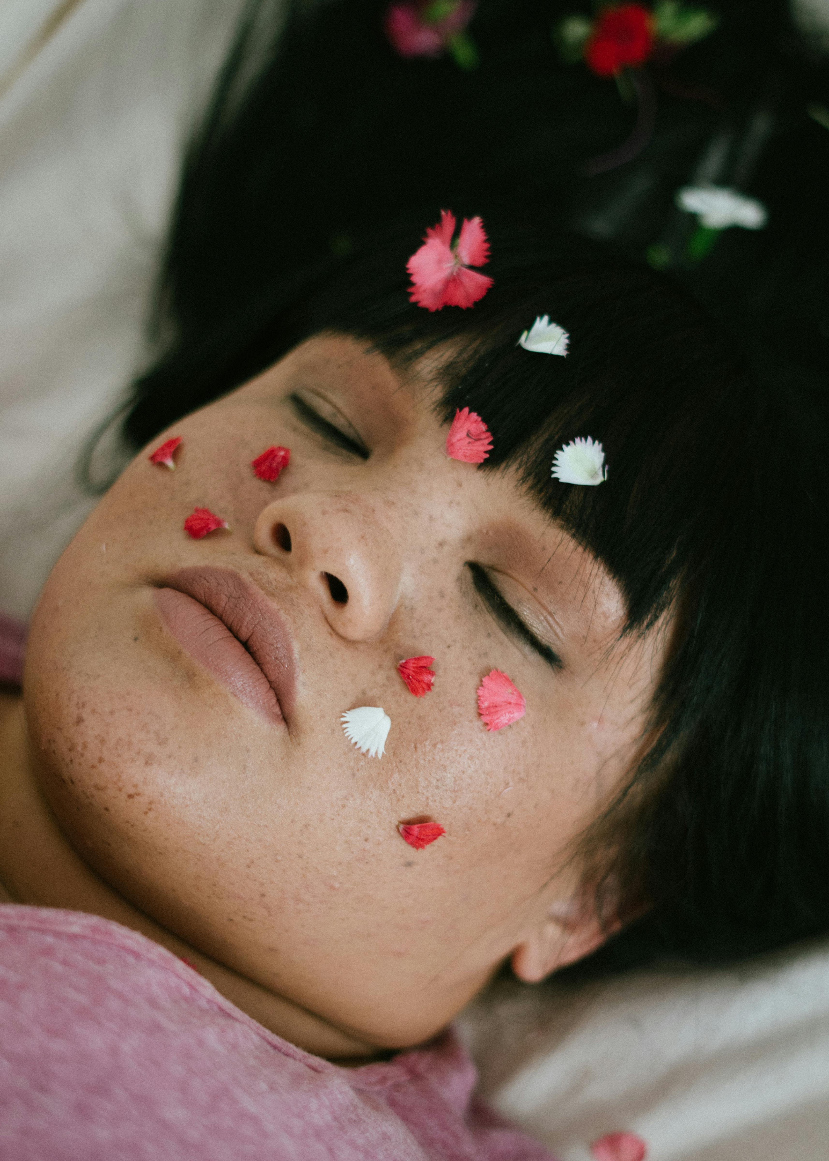 sensual plump ethnic lady relaxing on bed with flower petals on face and with closed eyes