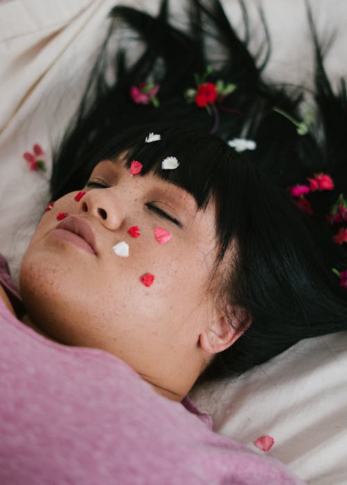 Sensual plump ethnic lady lying on bed with flower petals on face and with closed eyes