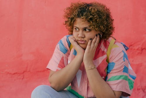 Thoughtful young ethnic plump lady in colorful clothes sitting in street near pink wall