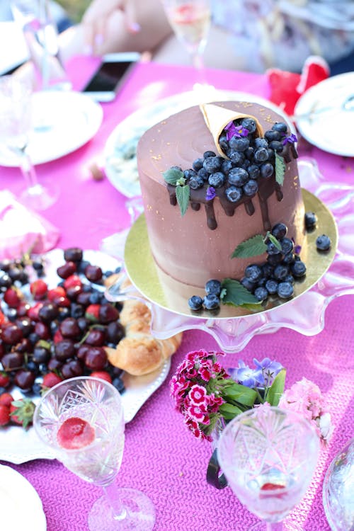 From above of chocolate cake with blueberry decoration and plate of berries on table in garden with glassware