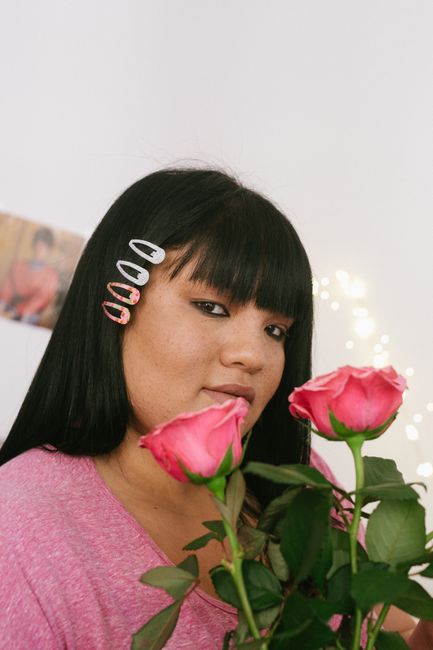 Thoughtful young ethnic overweight lady with black hair wearing pink shirt holding bouquet of pink roses while sitting in light room and looking at camera