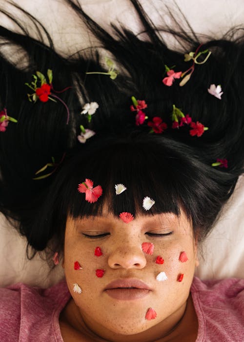 Free From above of young sensual ethnic plump lady with black hair and closed eyes wearing pink shirt with flower petals on face lying on white soft bed Stock Photo