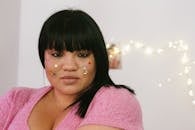 Tender young ethnic overweight lady with black hair and colourful glitter on face wearing pink shirt standing in light decorated room and looking at camera