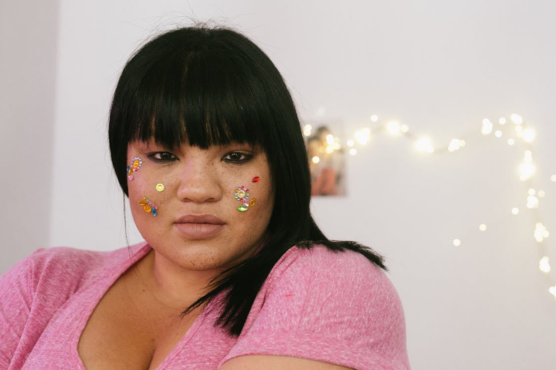 Tender young ethnic overweight lady with black hair and colourful glitter on face wearing pink shirt standing in light decorated room and looking at camera