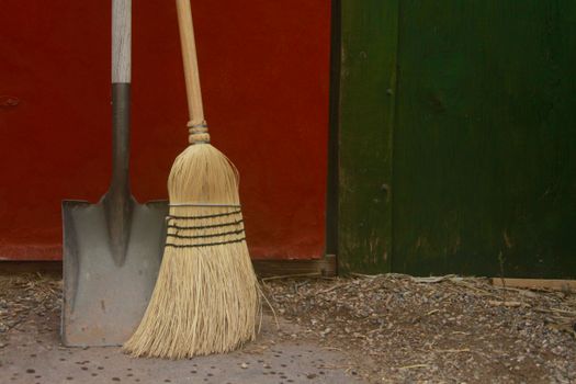 A rustic broom and shovel leaning against a red and green wooden shed outdoors.