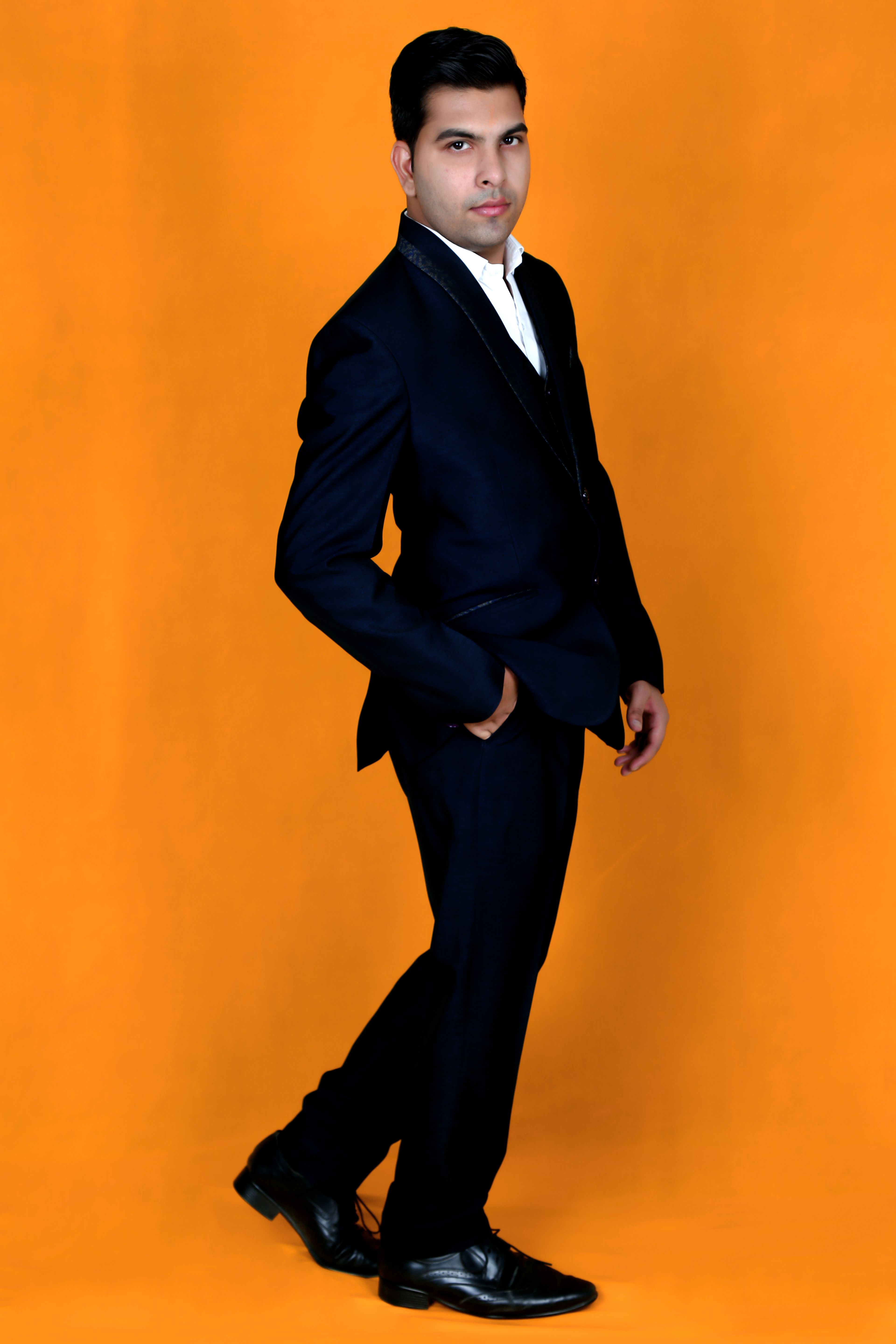 Portrait of a stylish young man in a black suit posing against an orange background.