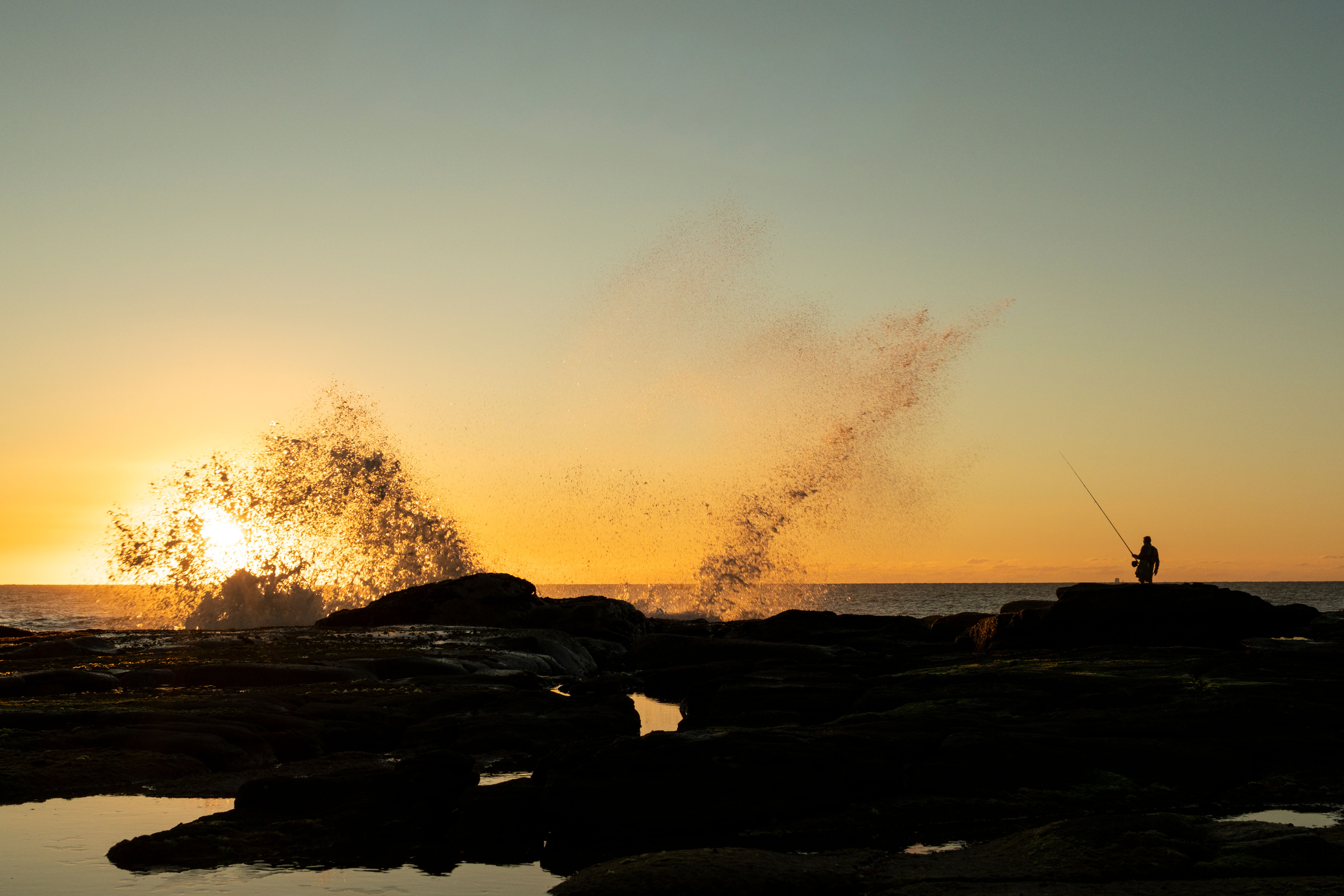Silhouette of a person fishing at sunrise with waves crashing on rocks, Sunshine Coast.