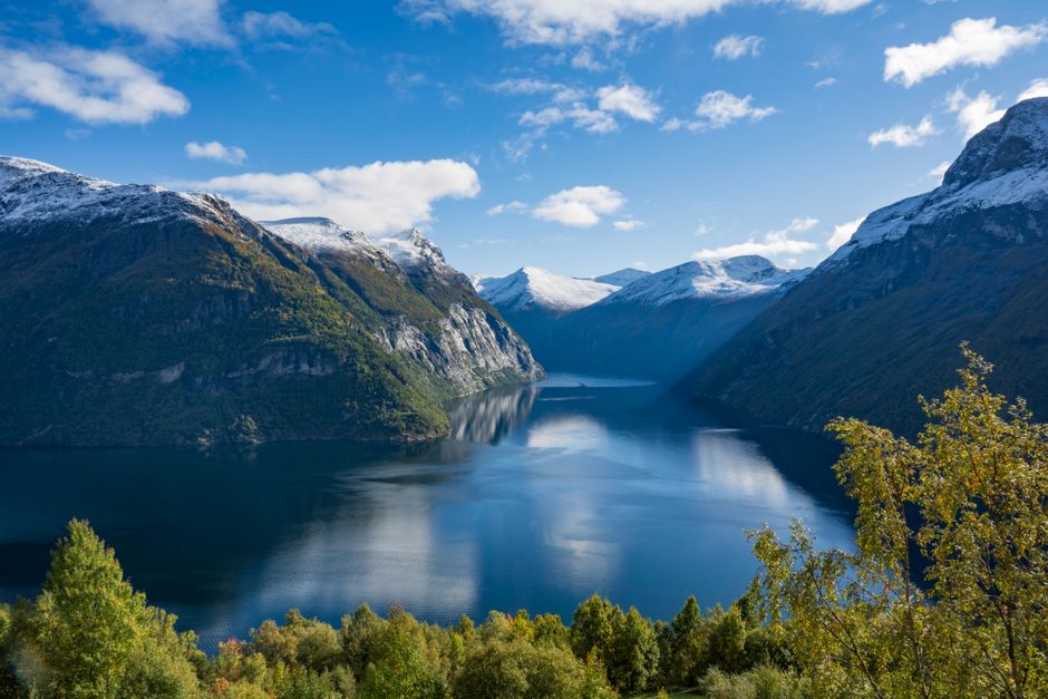 Breathtaking autumn view of a Norwegian fjord surrounded by snow-capped mountains and clear blue skies.