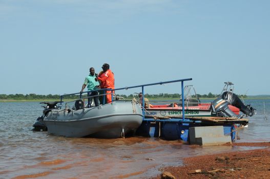 Two men on a boat discussing by a river bank in sunny Burkina Faso.